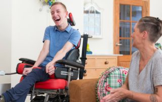 A young, physically impaired ALS patient in a red chair laughs with a caregiver in a grey shirt.