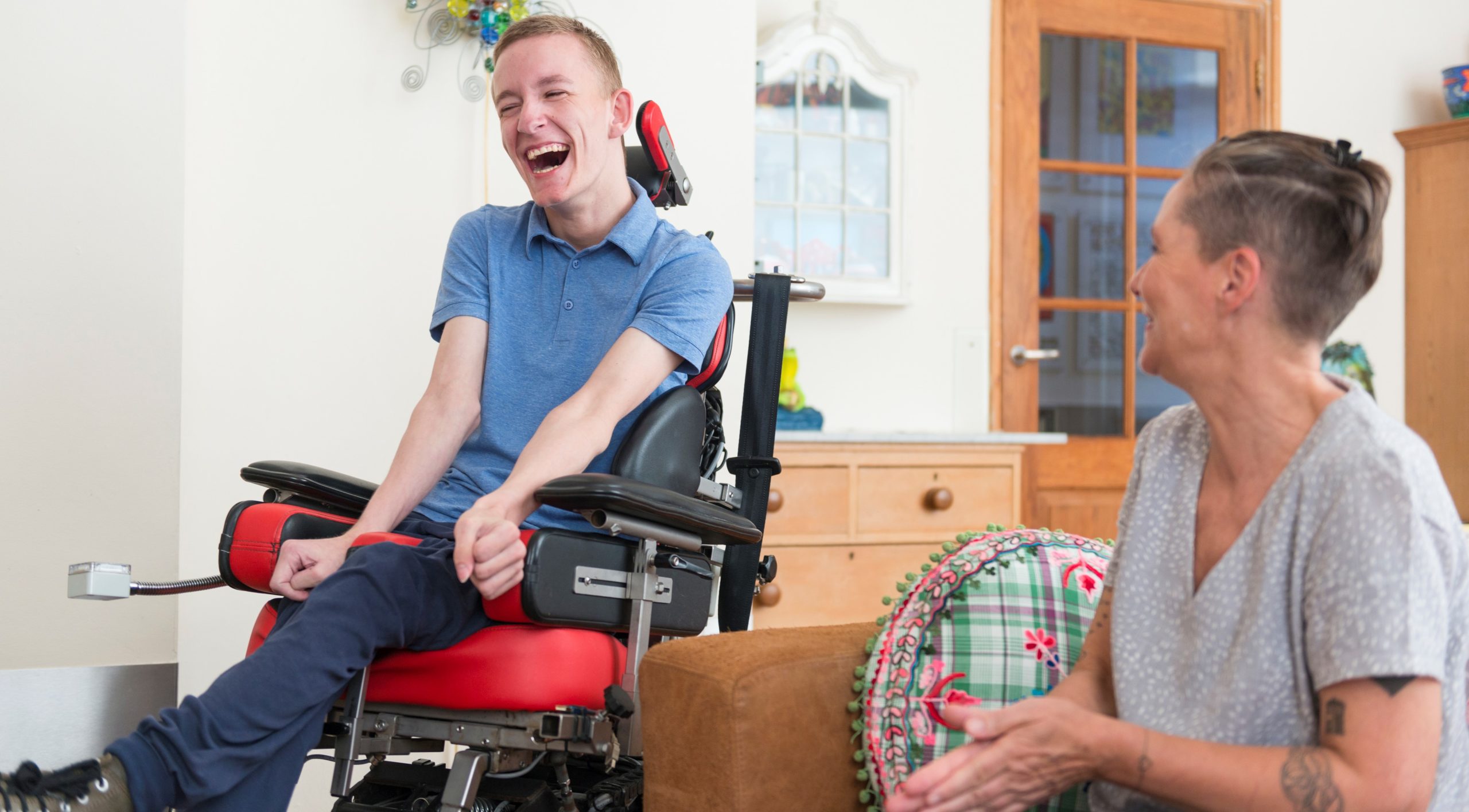A young, physically impaired ALS patient in a red chair laughs with a caregiver in a grey shirt.